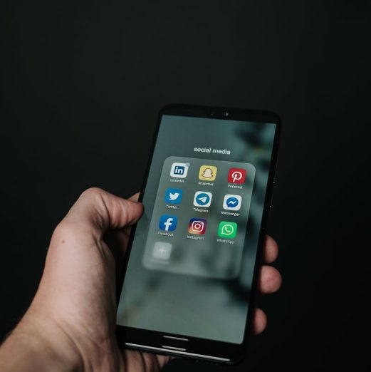 Close-up of a hand holding a smartphone displaying various social media app icons on a dark background.